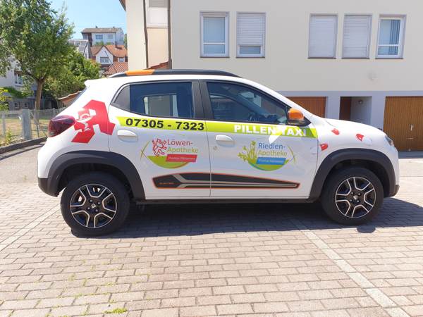 Ein Auto mit der Aufschrift „Pillentaxi“ der Riedlen-Apotheke in Ulm-Gögglingen steht auf einem Parkplatz. Das Fahrzeug zeigt die Logos der Riedlen-Apotheke und der Löwen-Apotheke.